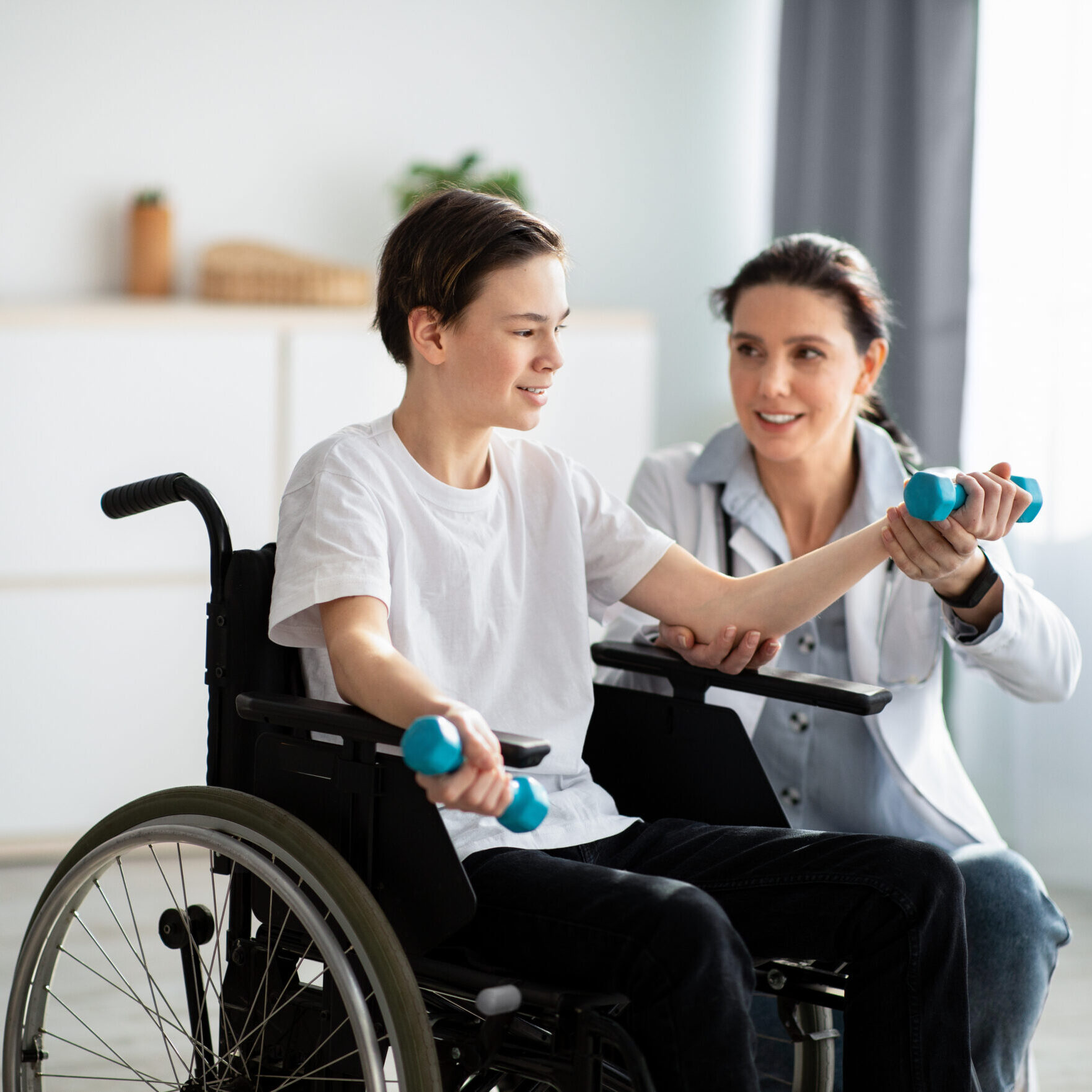 Physical rehabilitation concept. Young physiotherapist helping teenage boy in wheelchair to do exercises at home. Disabled adolescent training with dumbbells, working out his muscles indoors